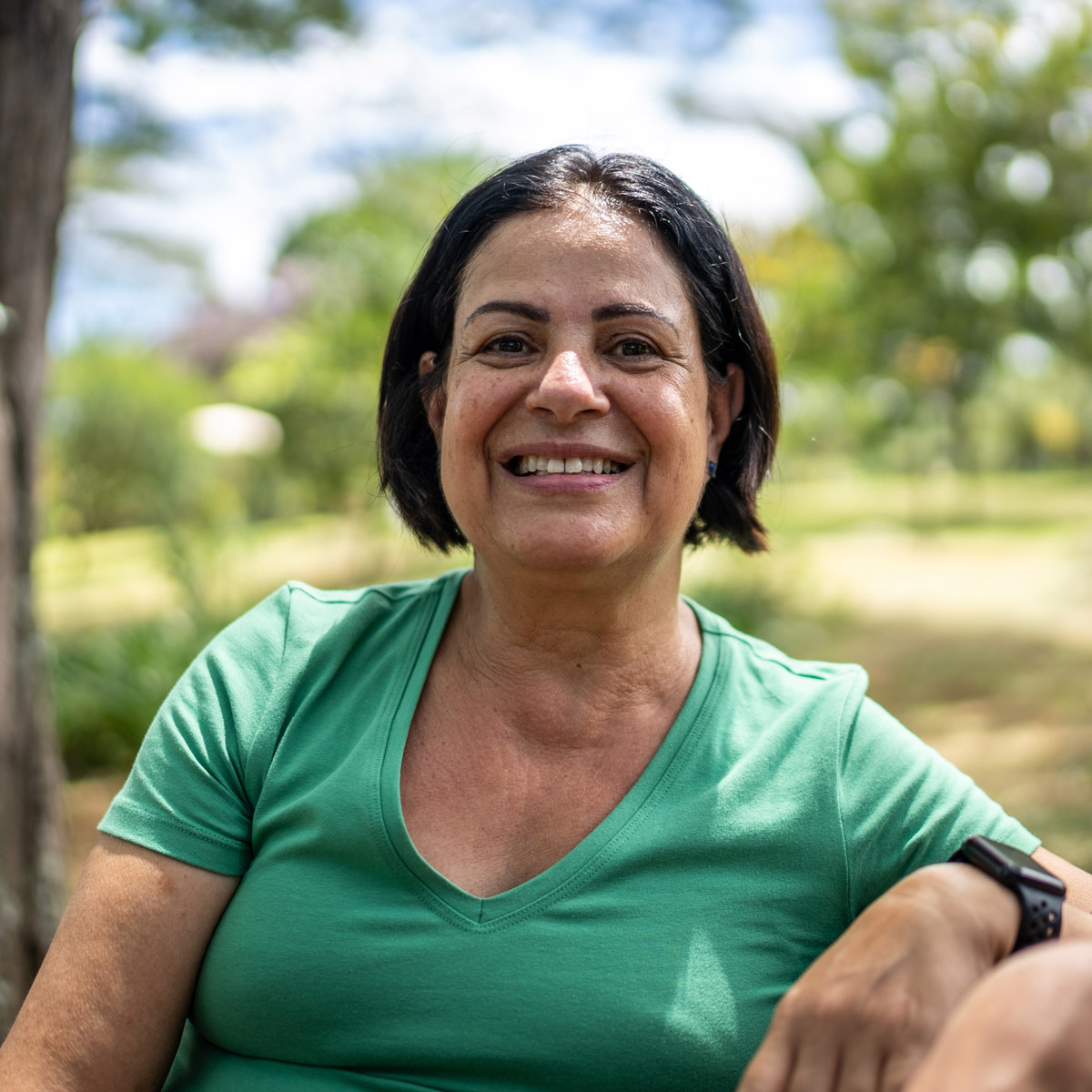 Smiling woman with short dark hair in a green t-shirt, sitting outdoors in a sunlit park.